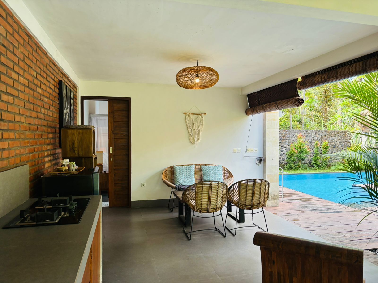Una habitación con pared de ladrillo, una mesa con sillas, una lámpara colgante y vista a una piscina.