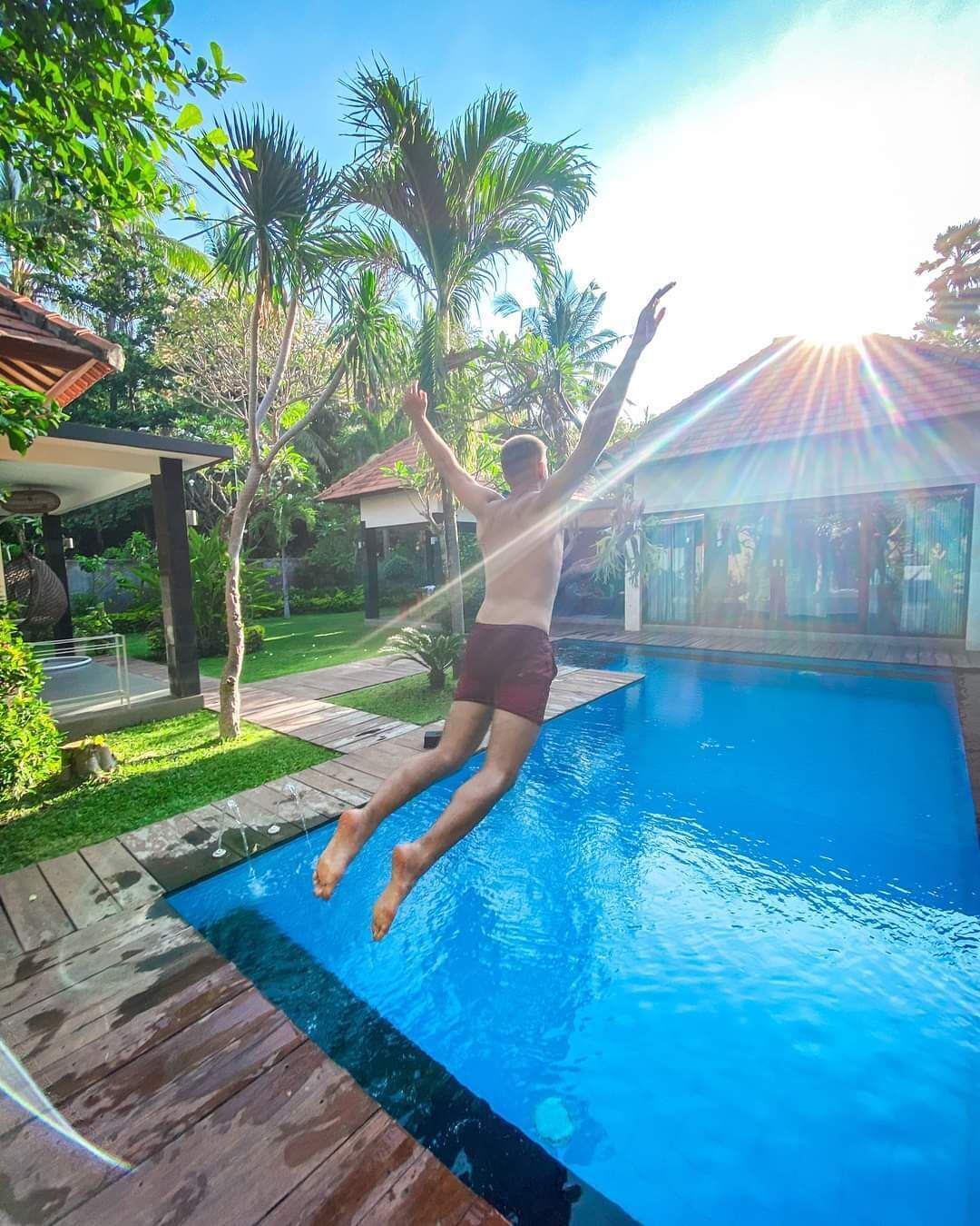 Une personne qui saute dans une piscine, avec une terrasse en bois et des palmiers en arrière-plan.