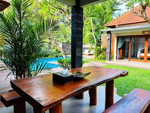 An outdoor wooden table with a bowl and napkin, surrounded by lush greenery and a swimming pool.