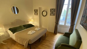 Ein Schlafzimmer mit einem Bett, einem Sessel, einem runden Spiegel und einem Fenster mit Vorhängen.