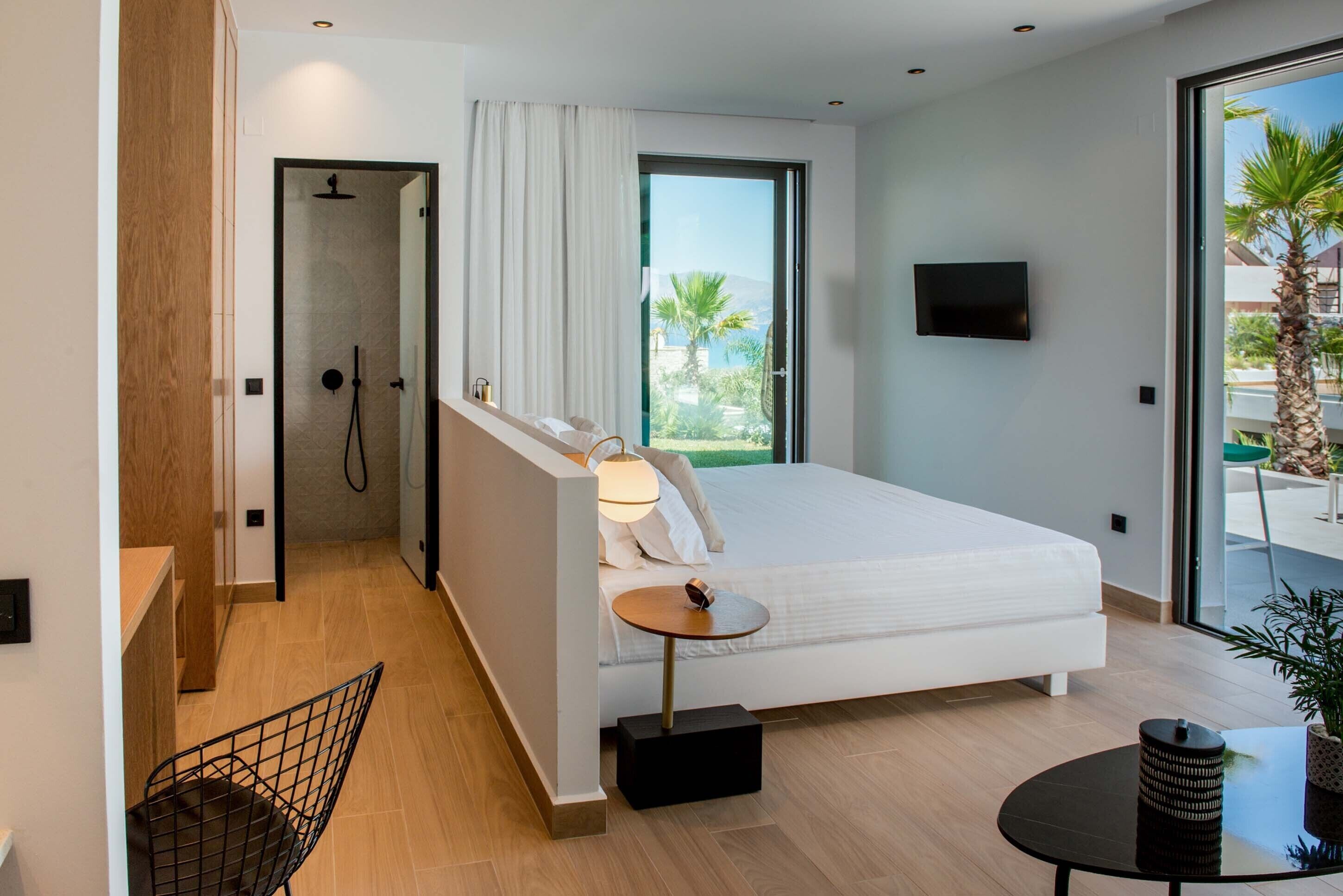 Quarto moderno com cama, uma mesinha, uma cadeira, televisão e área de chuveiro.