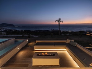 A rooftop terrace with a fire pit, outdoor seating, and a view of the ocean at dusk.