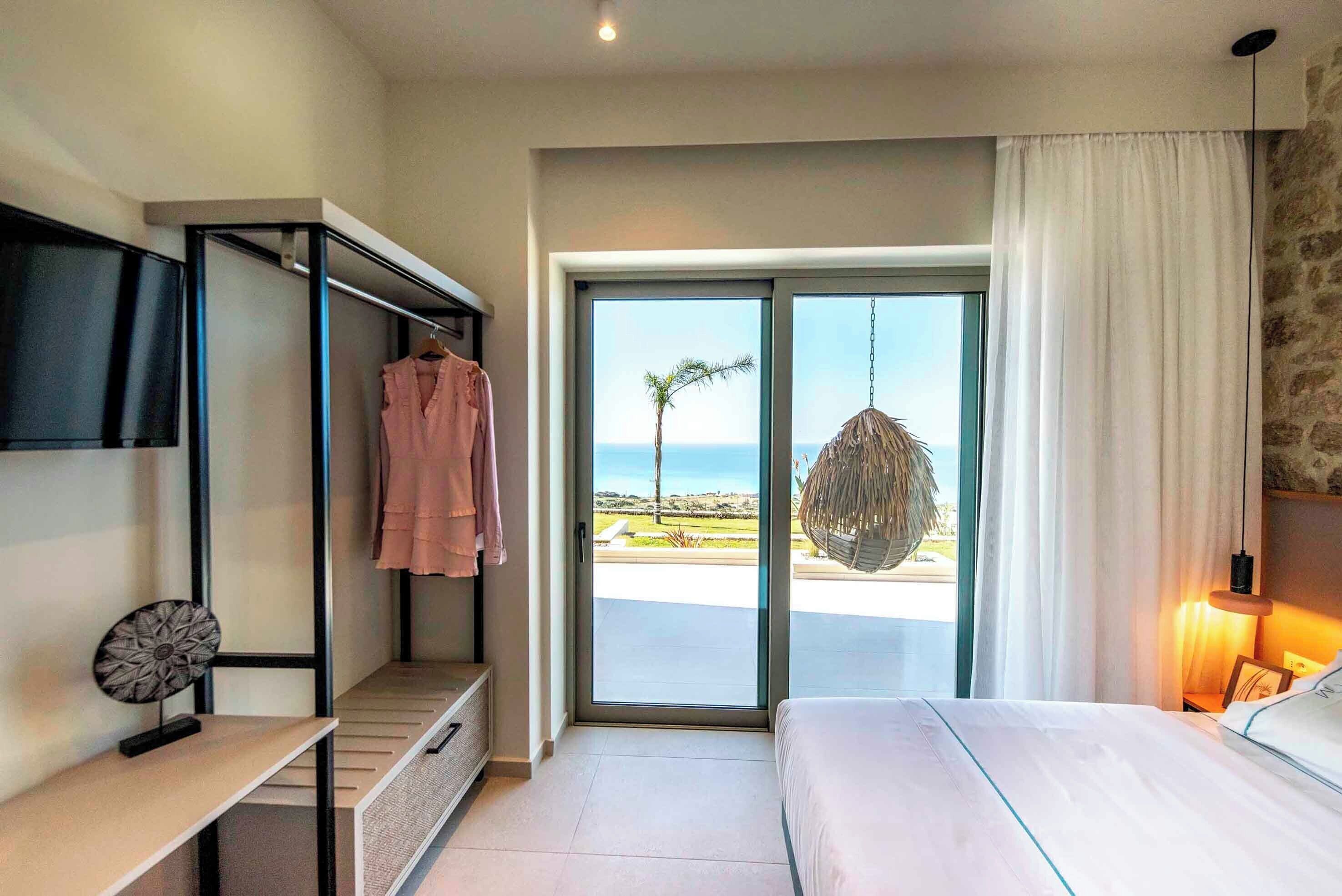 Een slaapkamer met een bed, een bureau, een hangstoel en uitzicht op het strand.