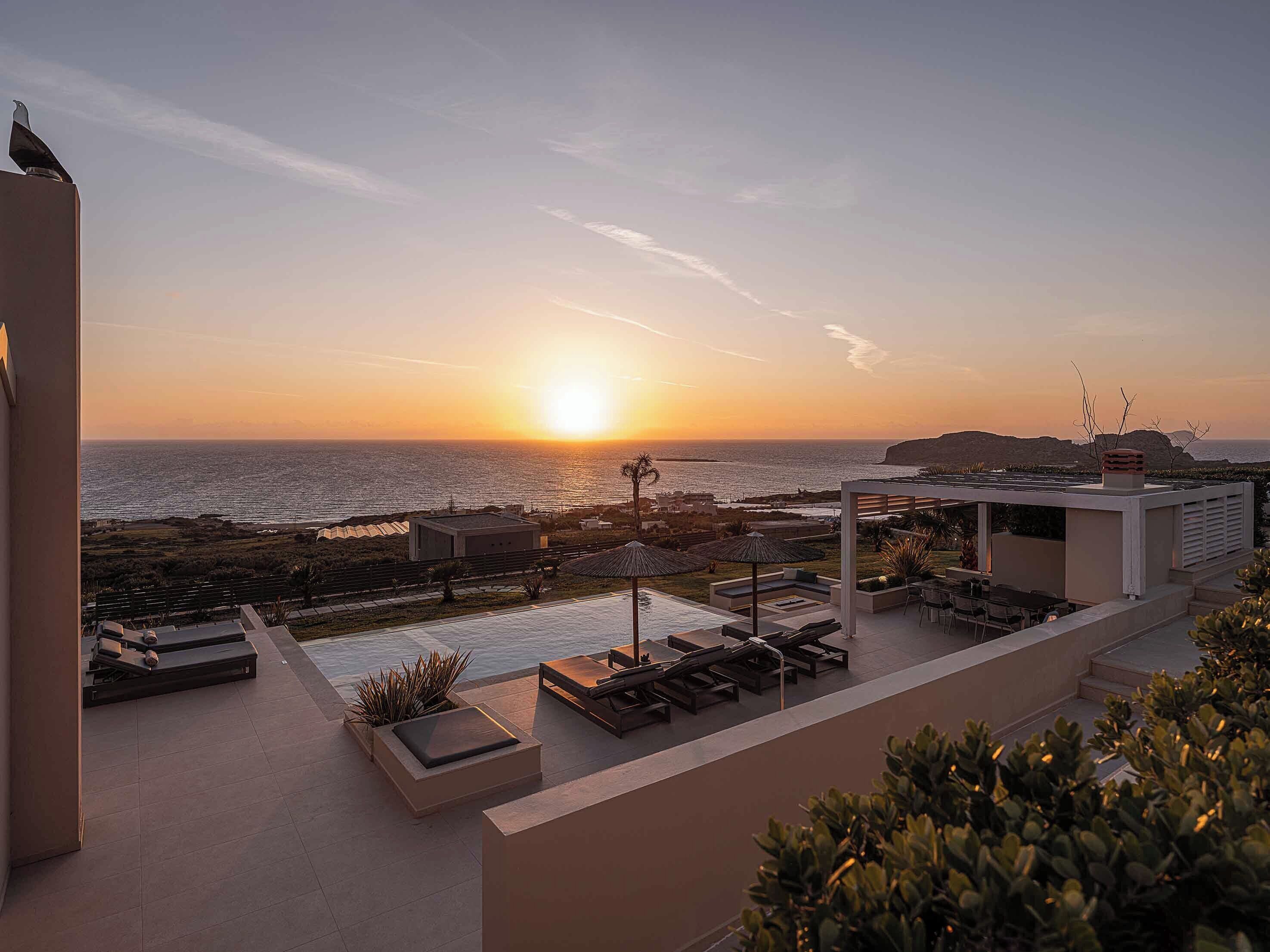 A rooftop terrace with a view of the sunset over the ocean, outdoor seating, and a pool area.