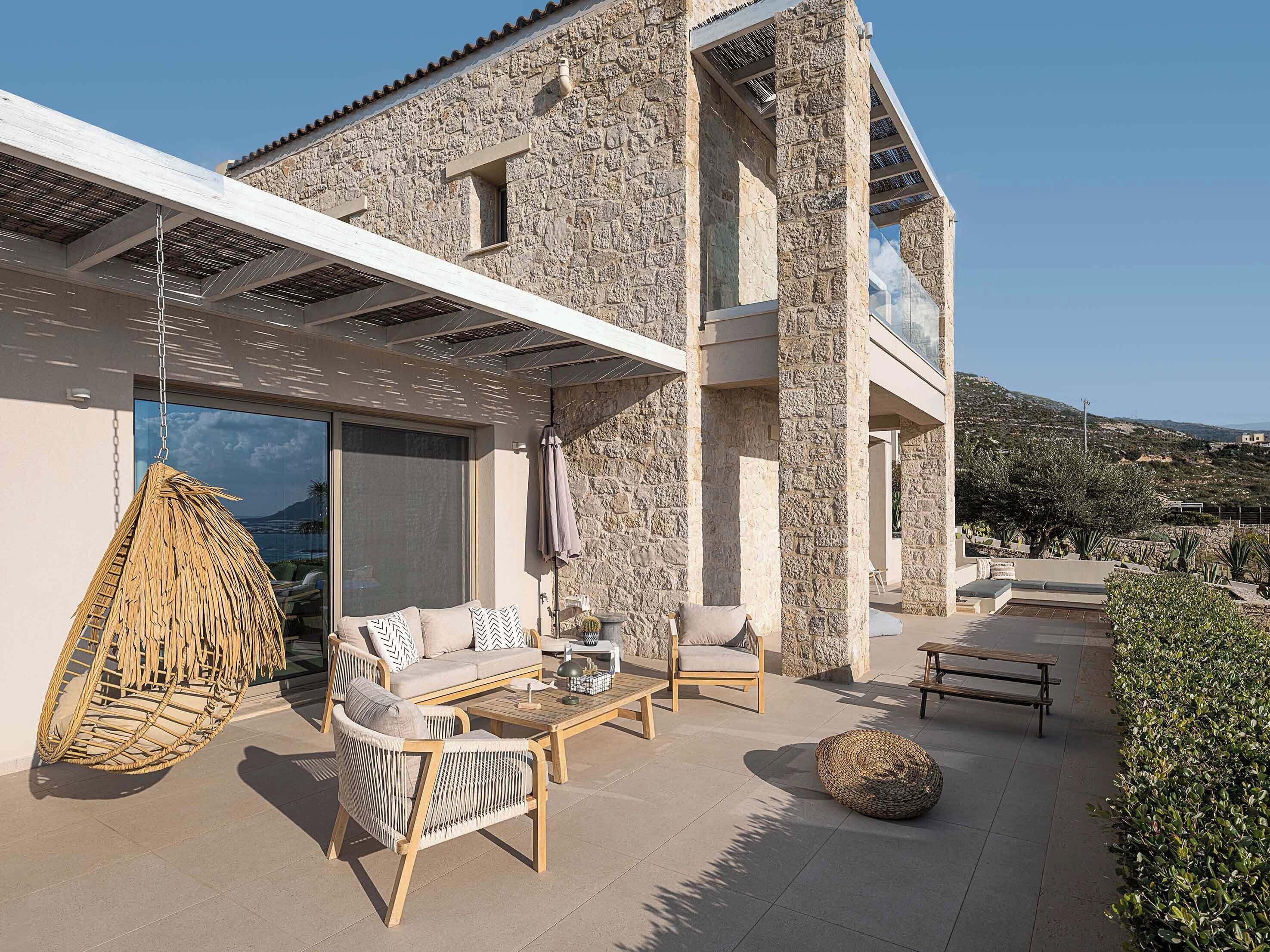 Una casa moderna con fachada de piedra, un porche cubierto con mobiliario y vistas del paisaje circundante.