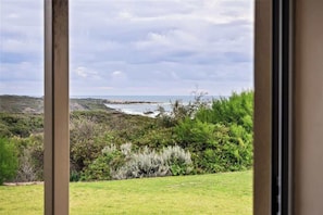 Eine Küstenlandschaft mit einer grasbewachsenen Vordergrundebene, dichter Strauchvegetation und Blick auf das Meer sowie ferne Hügel.