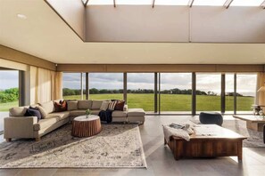 A spacious living room with a large sectional sofa, a round coffee table, and a view of a green field through large windows. - Incognito—An Exclusive Oceanfront Estate (Gracetown)