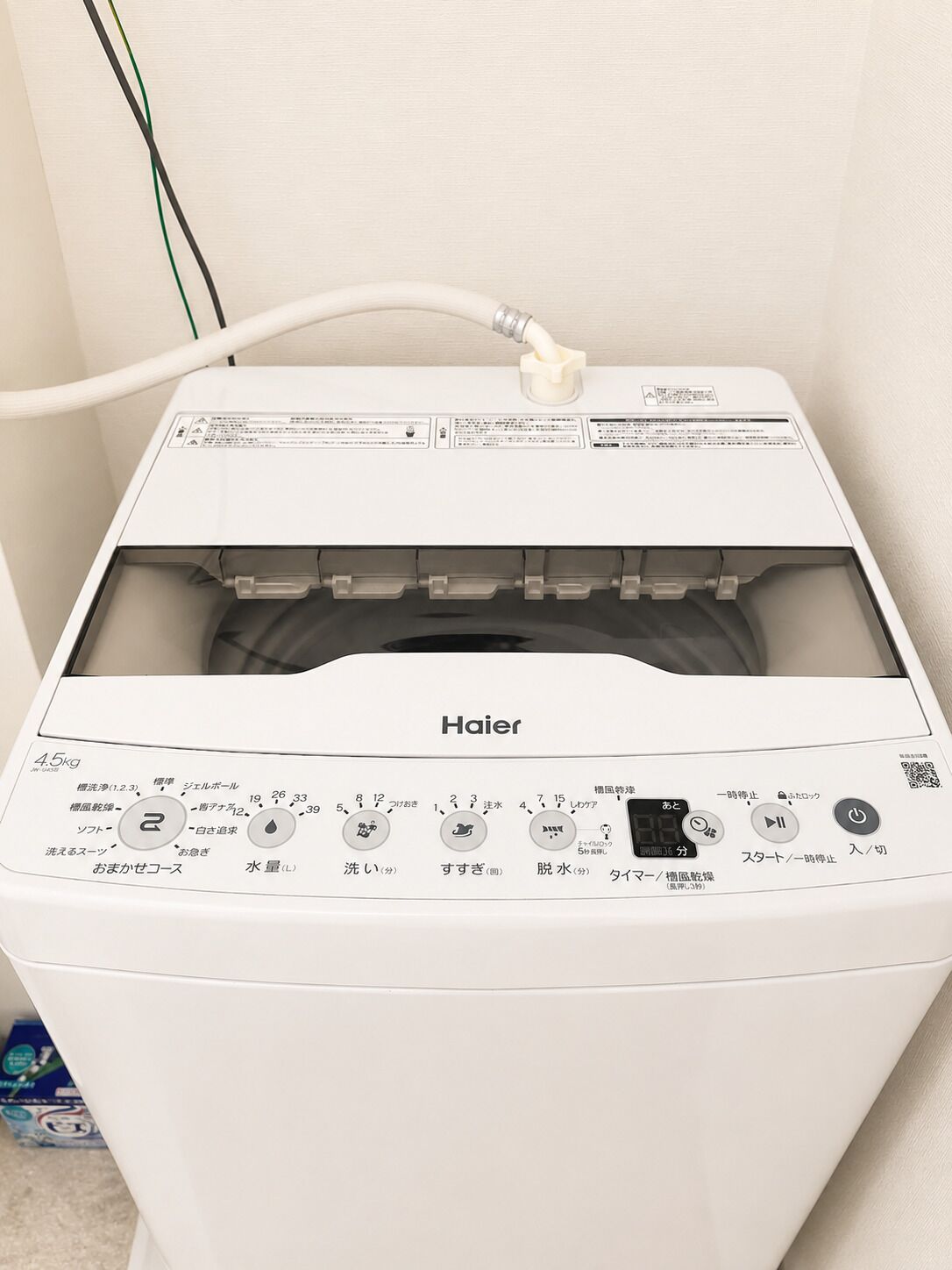 A white Haier washing machine with a control panel featuring multiple settings and a water inlet hose.