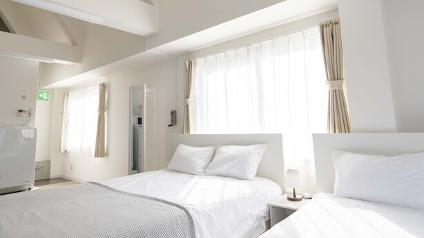 A hotel room with two beds, a ceiling fan, a window with curtains, and a bedside table. - ILIMA Higashi Nakano (Tokyo)