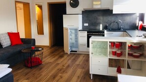 A compact living space with a kitchenette, a sofa with red cushions, a small table with a red bag, and a clock on the wall. - Vacation apartment "Fernblick " (ZWEIBRÜCKEN)