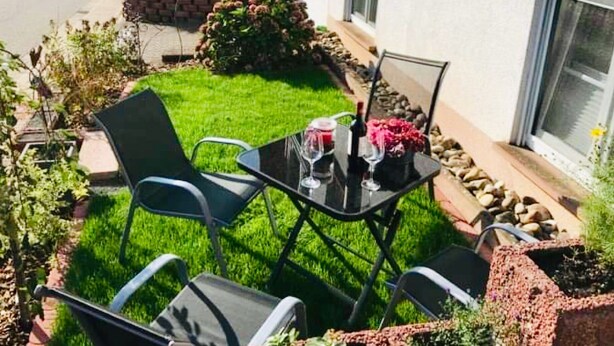 A patio with two chairs and a table, a small garden, and a white building with a brick wall and a window. - Vacation apartment "Fernblick " (ZWEIBRÜCKEN)