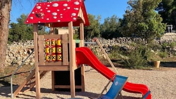 Une aire de jeux pour enfants avec un toboggan rouge et un mur d’escalade, entourée d’arbres et recouverte de gravier.
