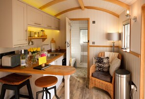 A compact kitchen with wooden cabinets, a built-in island with stools, a wicker chair, and a trash bin. - a beautiful rural shepherd hut with hot tub ! (Rushford)
