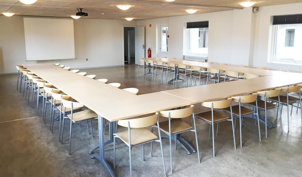 Un salón de conferencias con una larga mesa y sillas, una pantalla de proyector y ventanas.