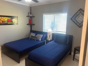 Quarto com duas camas de solteiro, um ventilador de teto, janela com persianas, uma luminária e um jogo de tabuleiro na parede.