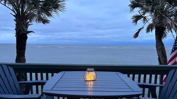 Una terraza con mesa y sillas, con vista al océano, palmeras y una bandera estadounidense.