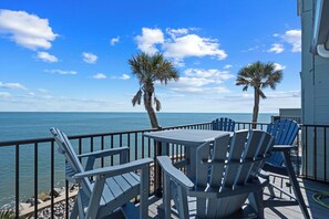 A deck with blue Adirondack chairs and a table, overlooking a calm sea with palm trees.