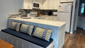 Eine moderne Küche mit weißen Schränken, einer eingebauten Mikrowelle, einem Kühlschrank und einer Sitzecke mit gepolstertem Sitz und gestreiften Kissen.