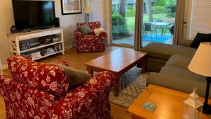 A living room with a TV, a sofa, a red floral armchair, a wooden coffee table, and a bookshelf.