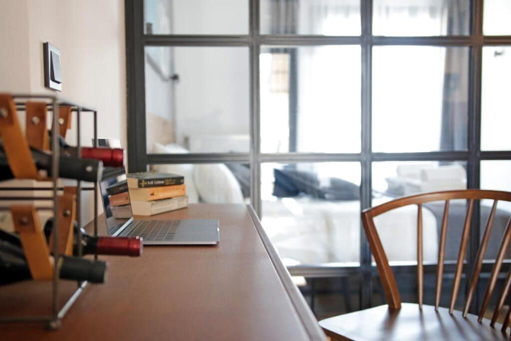 A wooden desk with a laptop, books, and a chair in a room with large windows.