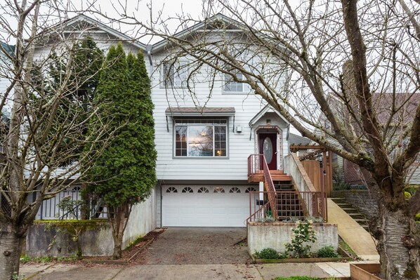 A two-story house with a front garage, a wooden staircase, and a small front yard with a tree.