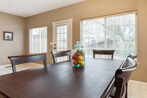 A dining area with a wooden table, chairs, a fruit jar, and large windows with blinds.