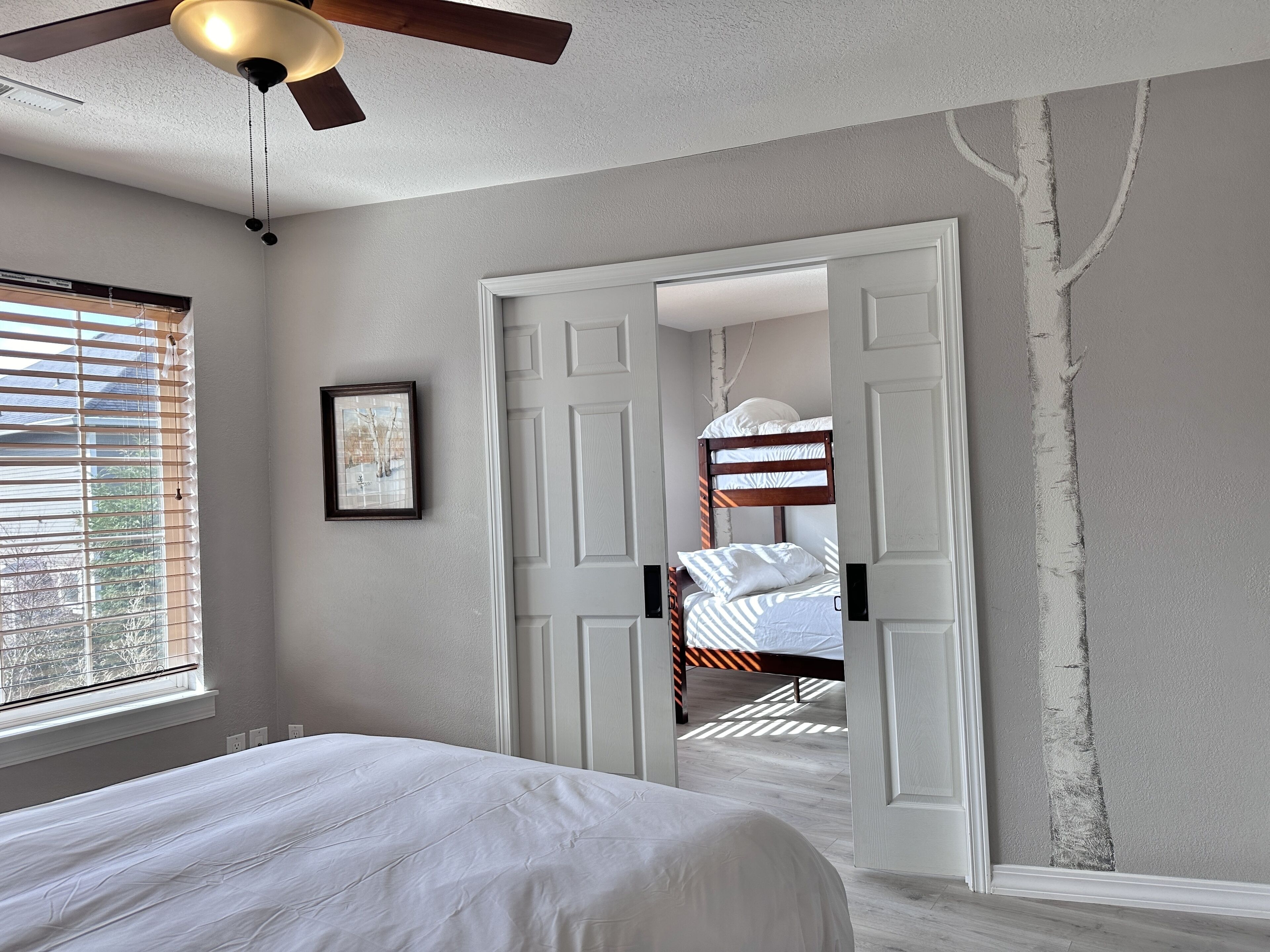 A bedroom with a bed, a ceiling fan, a window with blinds, and a door leading to another room.