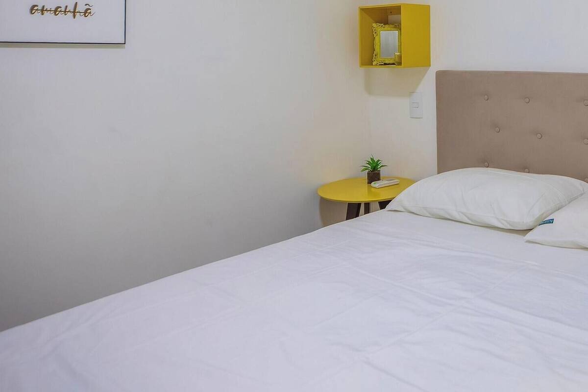 A neatly made bed with a tufted headboard, a small bedside table with a plant, and a yellow nightstand with a green object.