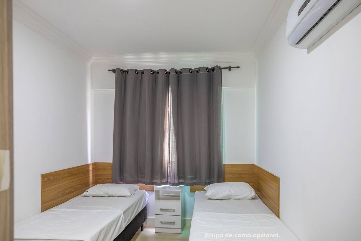 A small, clean room with two single beds, a wooden headboard, a bedside table, and a window with curtains.