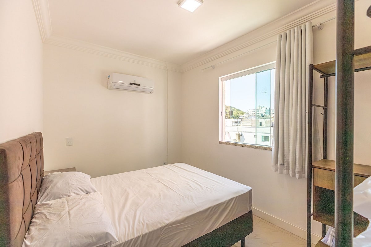 A bedroom with a single bed, a window with curtains, an air conditioning unit, and a view of buildings outside.