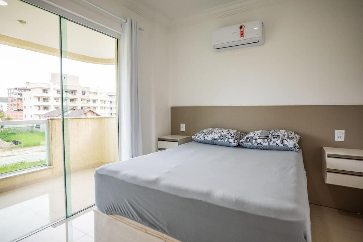 A bedroom with a bed, a nightstand, a wall-mounted air conditioner, and a view through a glass door.