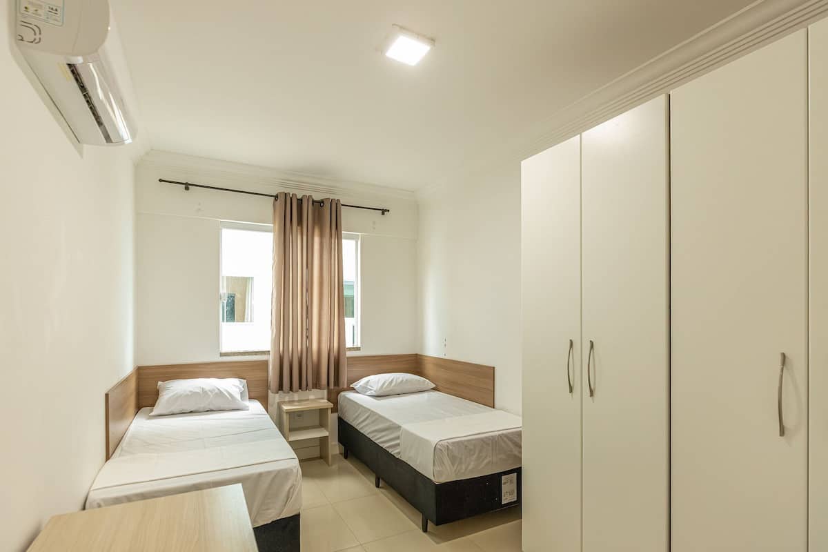 A small, clean room with two single beds, a nightstand, a wardrobe, and a window with curtains.
