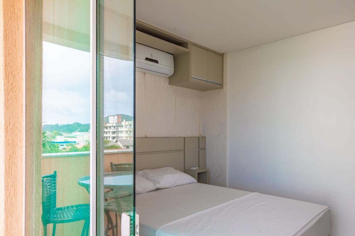 A hotel room with a bed, a small table, a chair, and a balcony with a view of buildings and greenery.