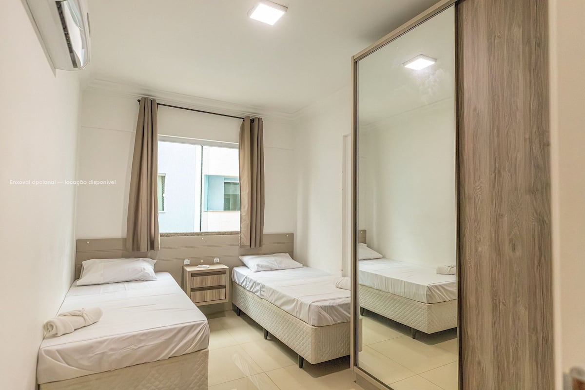 A hotel room with two beds, a large mirror, a window with curtains, and an air conditioning unit.