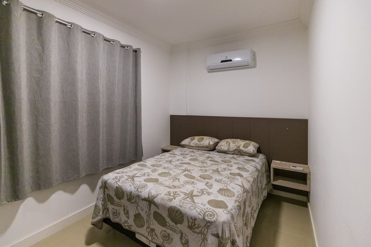 A bedroom with a bed, curtains, a nightstand, and an air conditioning unit.