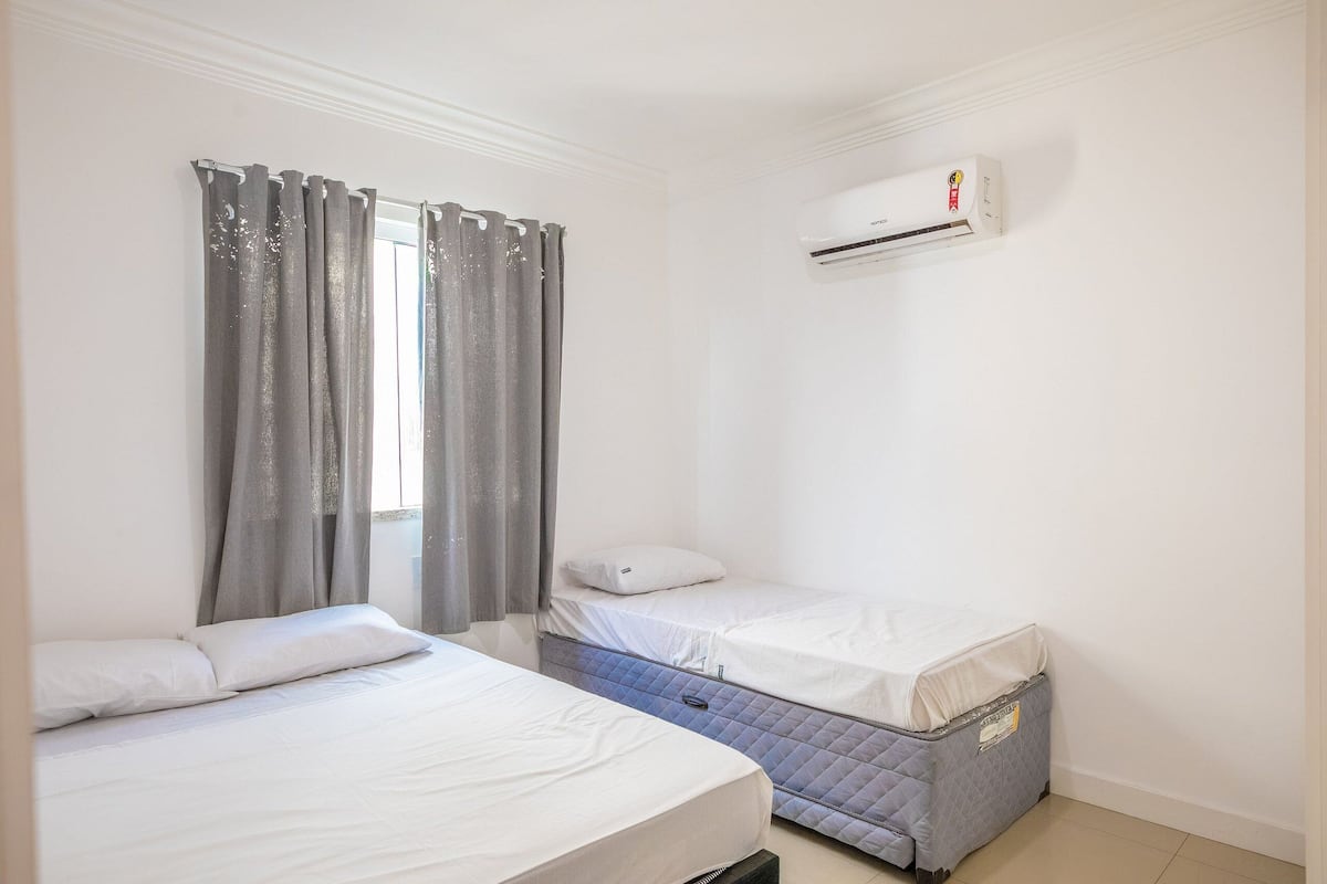 A small, clean room with two beds, a window with grey curtains, and an air conditioning unit on the wall.