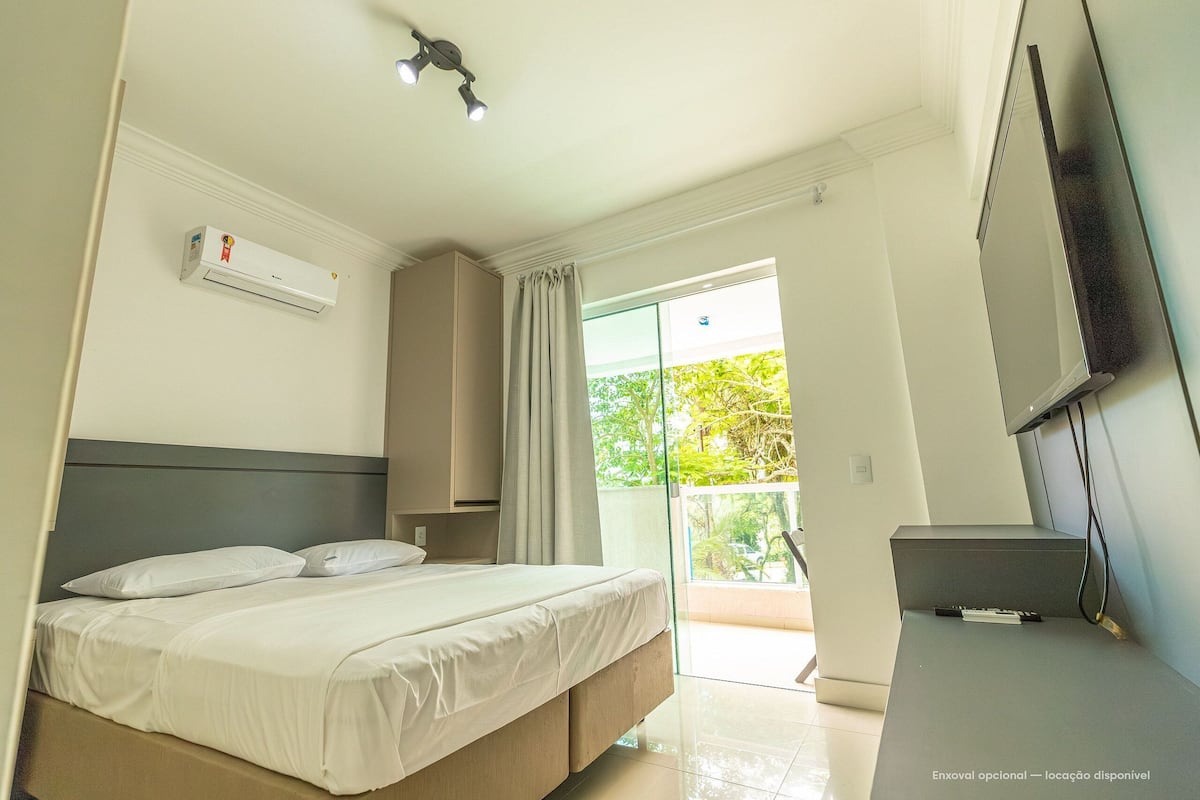 A bedroom with a bed, an air conditioning unit, a TV, and a balcony with a view of greenery.