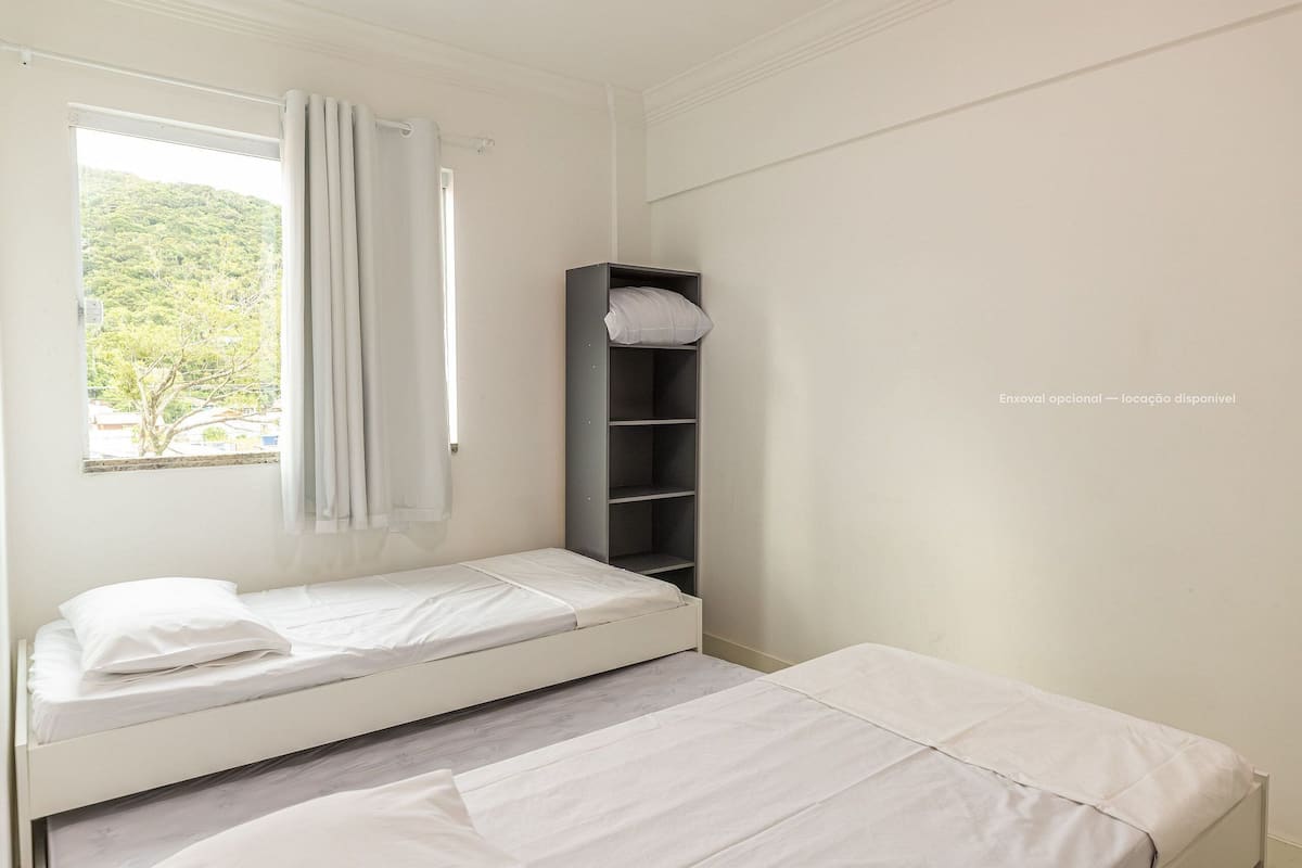 A small, clean room with two single beds, a window with curtains, and a built-in shelf with a pillow.