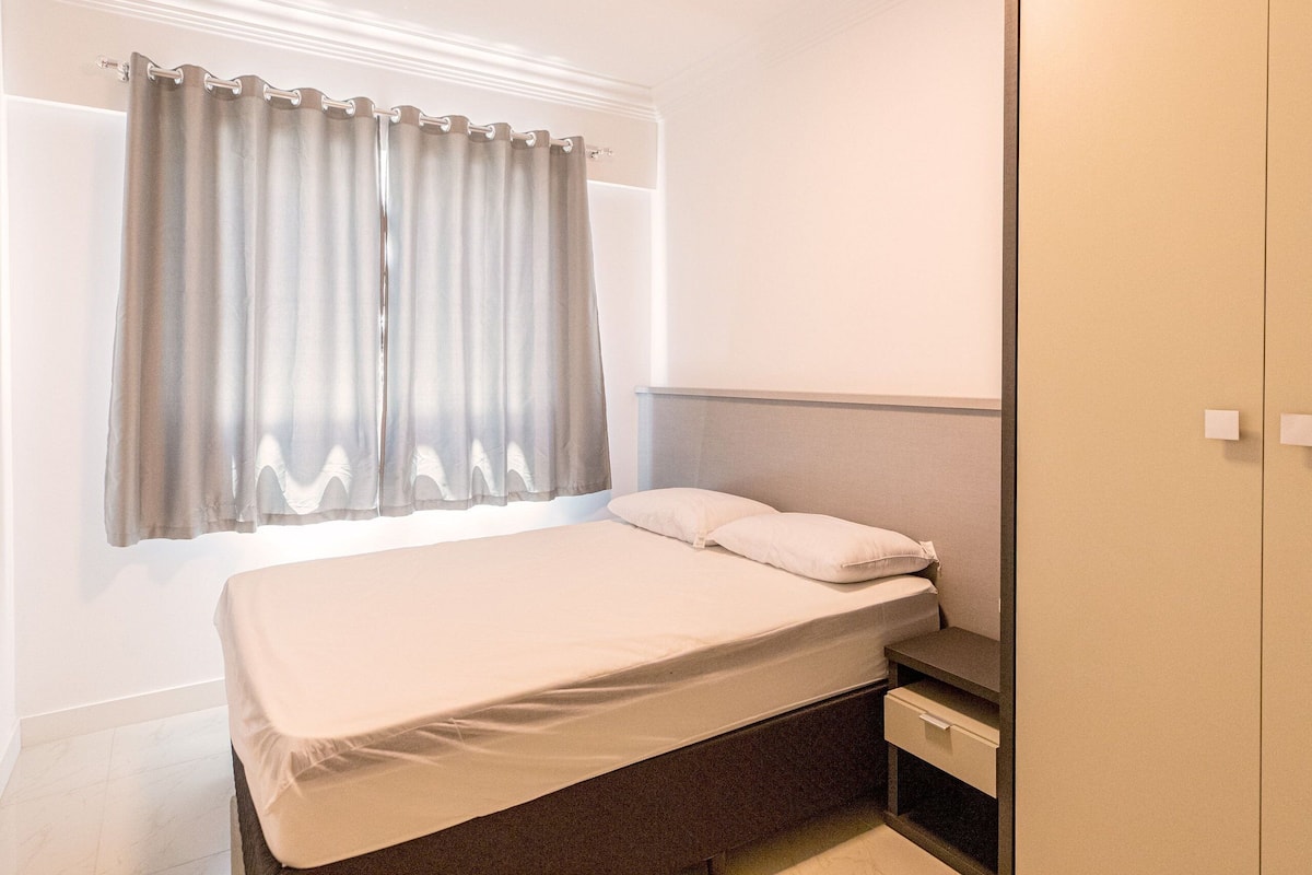 A bedroom with a bed, a nightstand, a drawer, and a window with curtains.