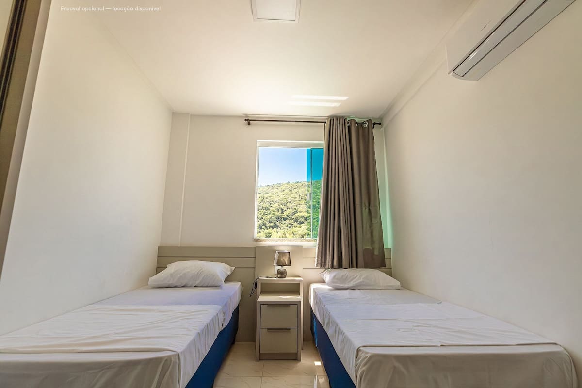 A small, clean room with two single beds, a nightstand with a lamp, and a window with curtains.