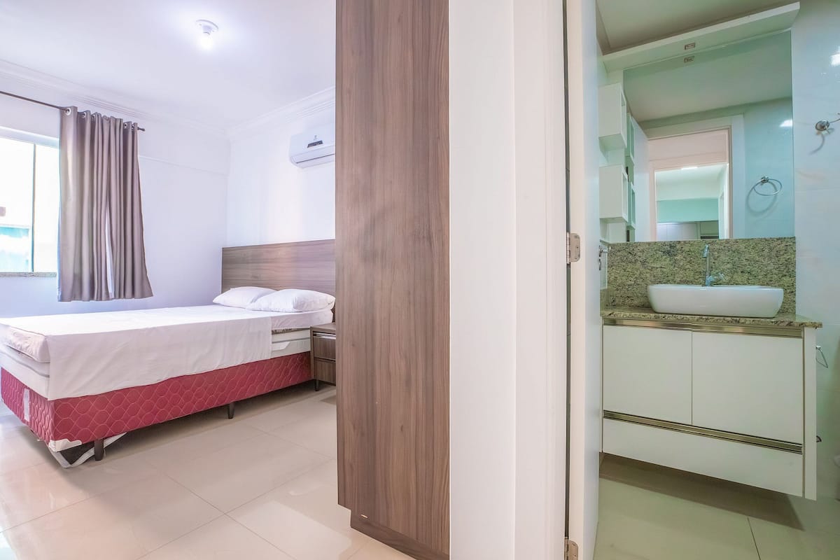 A hotel room with a bed, a wooden wardrobe, a bathroom with a sink and mirror, and a window with curtains.
