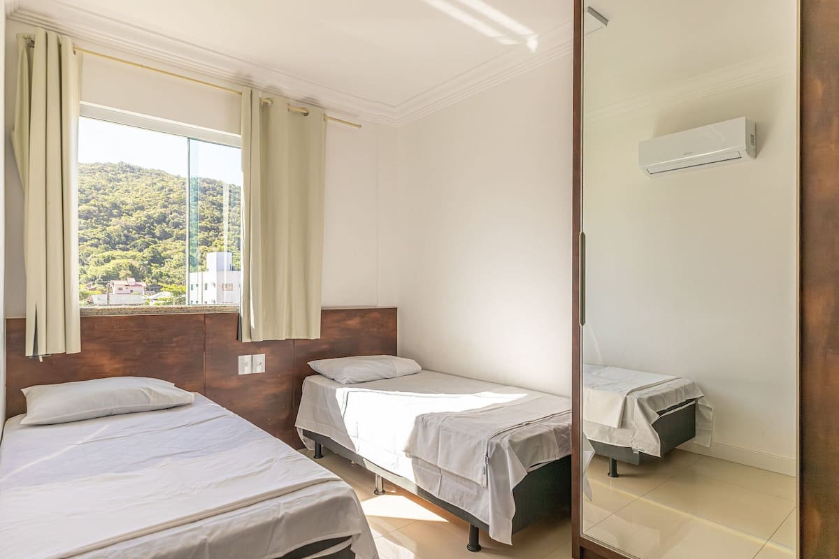 A hotel room with two beds, a large window with curtains, a mirror, and an air conditioning unit.