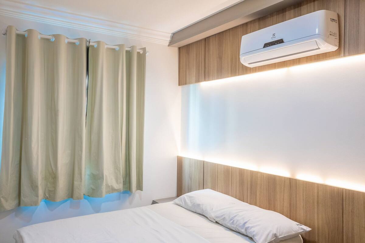 A hotel room with a bed, a wall-mounted air conditioner, and a window with curtains.