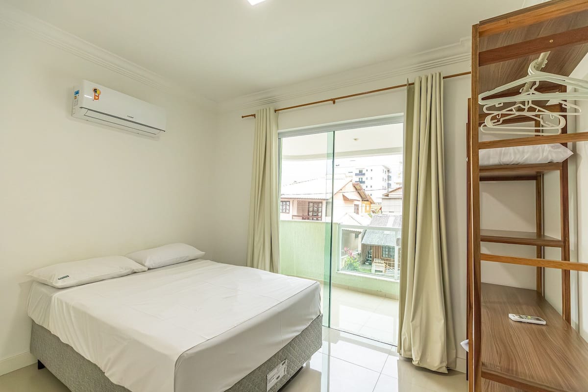 A bedroom with a bed, an air conditioning unit, a balcony with a view of buildings, and a wooden shelf with hangers.