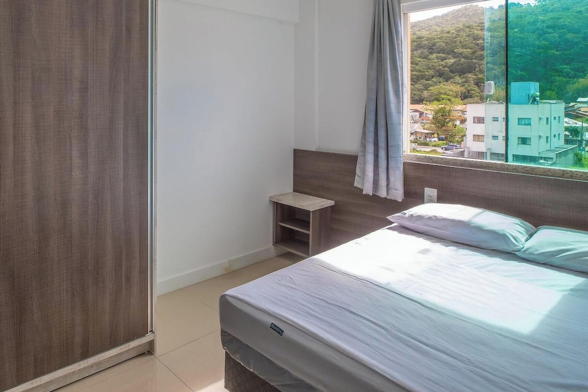 A bedroom with a bed, a nightstand, a wardrobe, and a window with a view of buildings and trees.