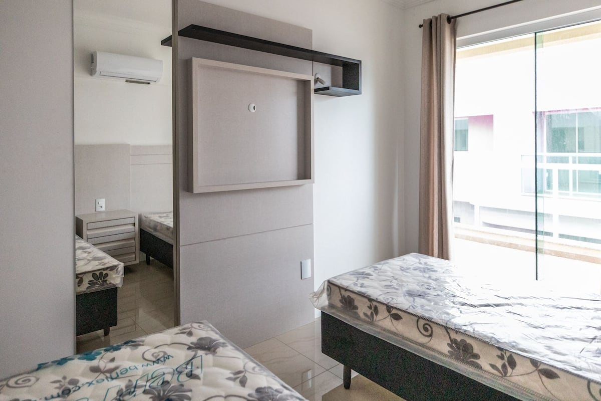 A bedroom with a bed, a small desk, a chair, a wardrobe, and a window with a view of buildings.