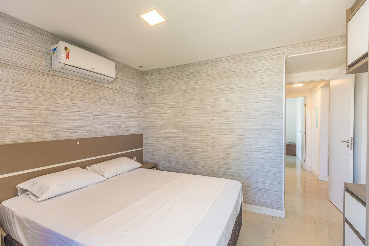 A hotel room with a large bed, a headboard, an air conditioning unit, and a view of a hallway.
