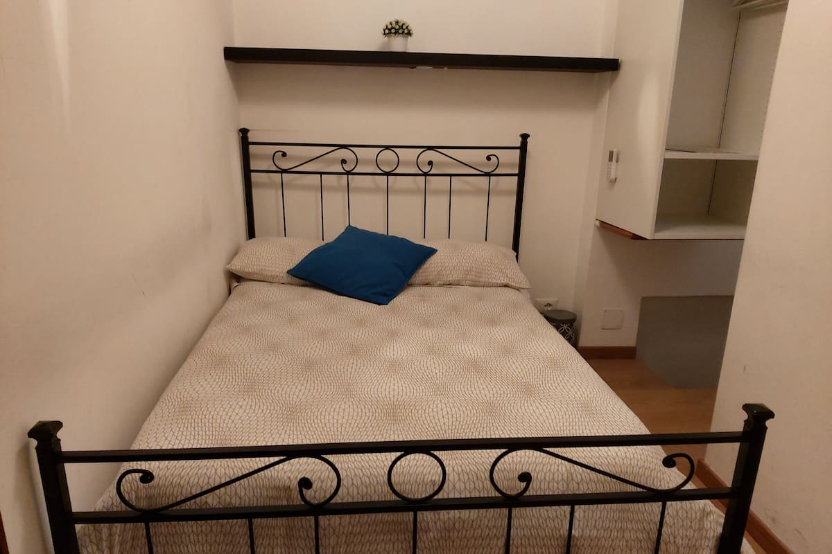 Ein kleines Zimmer mit einem Einzelbett, einem Metall-Kopfteil, einer gemusterten Bettdecke und einem blauen Kopfkissen.