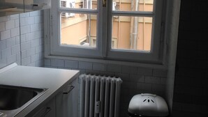 Ett litet kök med ett fönster, vita skåp, en diskho, ett element och en papperskorg.