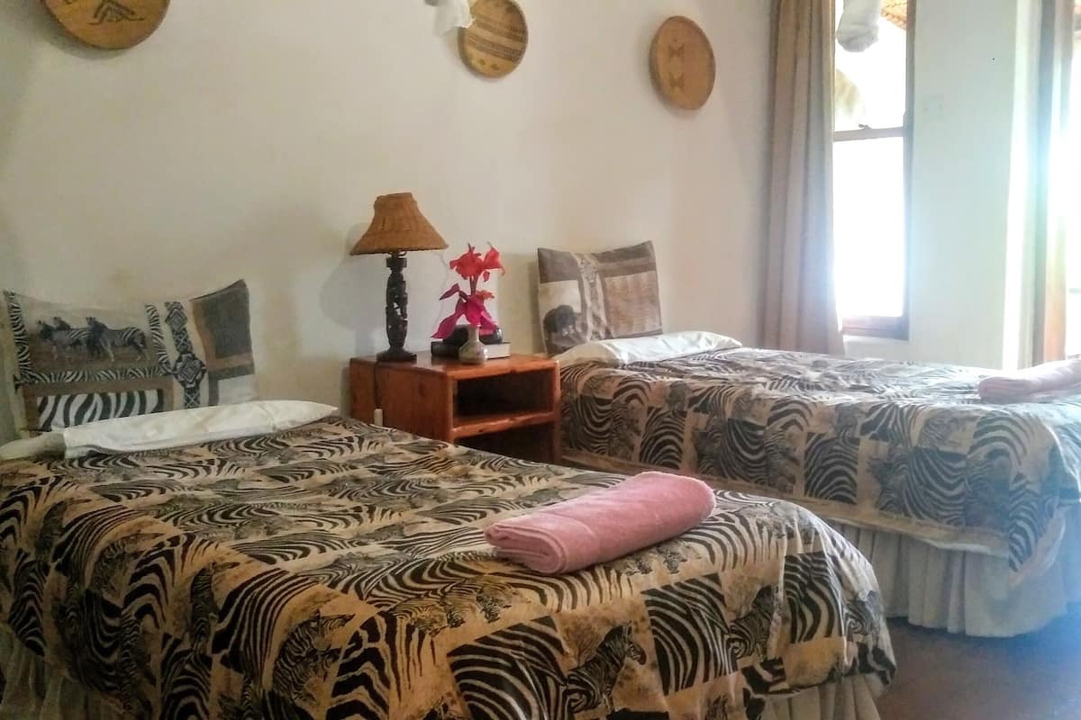 Un dormitorio con dos camas, una mesita de noche, una lámpara, almohadas decorativas y una ventana con cortinas.